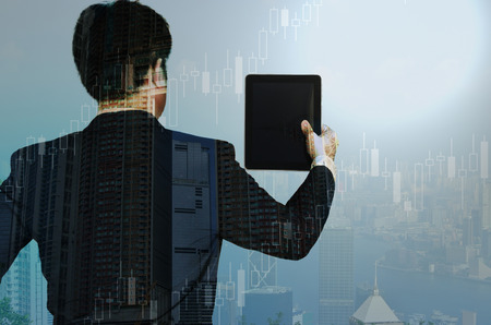 Double exposure of businessman using tablet to manage his business with financial graph chart and cityscape blurred building background. business vision conceptの写真素材