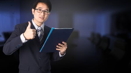 Confident Asian businessman showing document file and giving thumbs up in front of meeting room, looking at camera, smiling.の写真素材