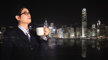 Double exposure of businessman holding cup of coffee looking and thinking with cityscape night scene blurred building background.の写真素材