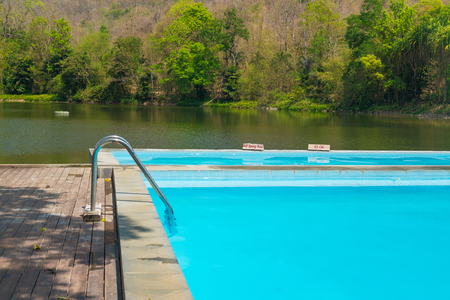 Blue swimming pool with wooden flooring near the lake mountain and tree with blue sky and cloud. under sunlight.の写真素材
