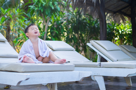 Asian boy with white towel resting on a lounge deck chair or sun lounger near swimming pool with big tree and sun light. on summer holidays.の写真素材