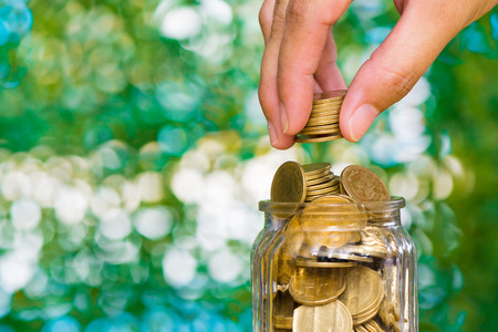 Woman hand put gold coin money in the glass jar on table in garden with green background, for saving for the future banking finance business concept.の写真素材