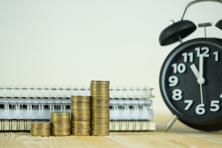 Steps of coins stack with vintage alarm clock and pen, notebook paper on wooden working table with...