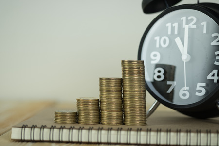 Steps of coins stack with vintage alarm clock and pen, notebook paper on wooden working table with...