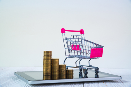 Step of coin stack and shopping cart or supermarket trolley on tablet computer screen on white working table, business and finance concept idea.の写真素材