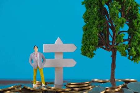 Figure miniature businessman or small people investor standing on coin stack with white wooden board...