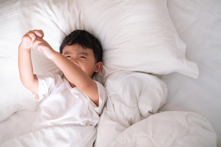 3 years old little cute Asian boy in white shirt at home on the bed, kid lying playing and smiling on white bed with pillow and blanket, top view with copy space for add text.の写真素材