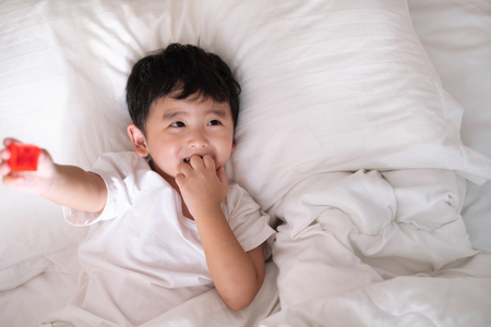 3 years old little cute Asian boy in white shirt at home on the bed, kid lying playing and smiling on white bed with pillow and blanket, top view with copy space for add text.の写真素材