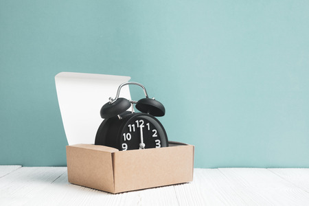 Retro alarm clock in brown delivery cardboard box or tray on white wood and blue wall background,...