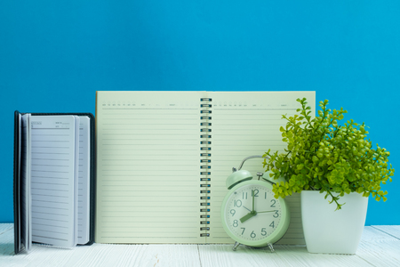 Office supplies or office work essential tools items on wooden desk in workplace, notebook, alarm clock and little tree with blue wall background copy spaceの写真素材