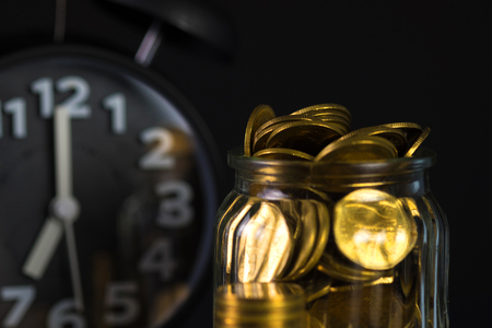 Coins stacks with coin in glass jar bottle and alarm clock in dark room, business and finance concept idea.の写真素材