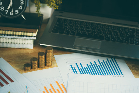 Coin stack and financial graph paper sheet with notebook calculator and alarm clock on working table, business finance concept idea.の写真素材