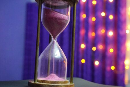 a sand clock with light purple sand with bokeh background and dark purple curtainsの写真素材