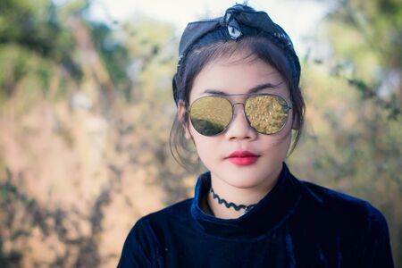 Fashion pretty young woman model in hipster style,fashion black glasses,stands over forest background. Outdoor portrait of cute girl having fun.の写真素材