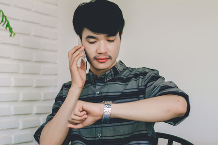 A young man looking at the wrist watch while talking on a cell phone.の写真素材