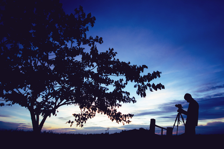 Silhouette photographer with camera on tripod taking pictures of nature tree on sunset.の写真素材