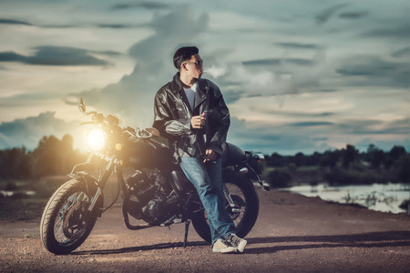 Biker man standing smokes with his motorbike beside the natural lake and beautiful, enjoying freedom and active lifestyle, having fun on a bikers tour.Night background and sky.の写真素材