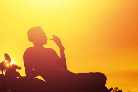 Silhouette of biker man sitting smokes with his motorbike , enjoying freedom and active lifestyle, having fun on a bikers tour.sunset background and sky.の写真素材