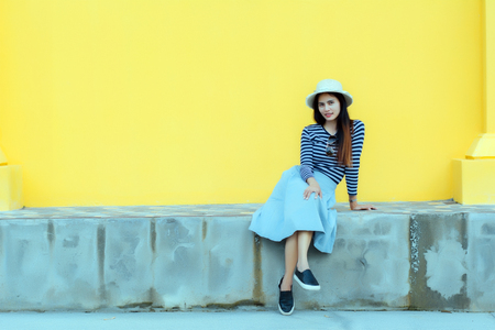 Sunny lifestyle fashion portrait of young stylish hipster woman beside building against yellow background, wearing trendy outfit, hat, travel concept.の写真素材
