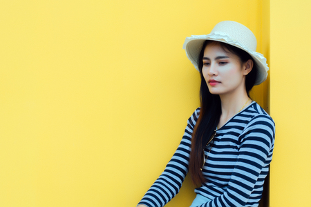 Sunny lifestyle fashion portrait of young stylish hipster woman beside building against yellow background, wearing trendy outfit, hat, travel concept.の写真素材