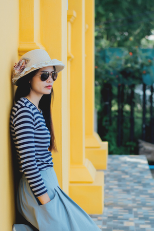 Sunny lifestyle fashion portrait of young stylish hipster woman beside building against yellow background, wearing trendy outfit, hat, travel concept.の写真素材