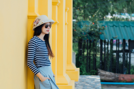 Sunny lifestyle fashion portrait of young stylish hipster woman beside building against yellow background, wearing trendy outfit, hat, travel concept.の写真素材