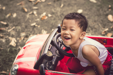 Little boy driving car with the steering wheel. Boy in a white shirt in a red toy car in the street. Little boy driving big toy car and having fun, outdoors.の写真素材