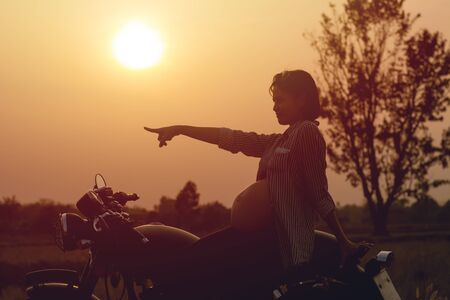 Cose-up of biker woman a pregnant woman on a motorcycle on sunset background. She holding a flower grass. Tip and lifestyle of motorbike concept.の写真素材