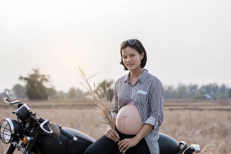 Cose-up of biker woman a pregnant woman on a motorcycle on sunset background. She holding a flower grass. Tip and lifestyle of motorbike concept.の写真素材
