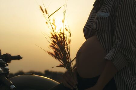Cose-up of biker woman a pregnant woman on a motorcycle on sunset background. She holding a flower grass. Tip and lifestyle of motorbike concept.の写真素材