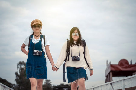 Two trendy cool hipster girls, friends, on the old wooden bridge, and backpacks, holding vintage camera, positive emotions.の写真素材