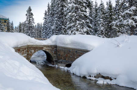 Winter landscape in the mountains with lots of white fresh snow, covered in snow trees, an unfrozen creek and a stone bridgeの写真素材