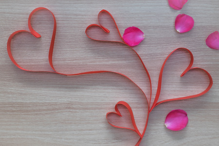Four red heart shape ribbon with pink rose petals on wooden surface for valentines or marriage proposal with empty space for textの写真素材