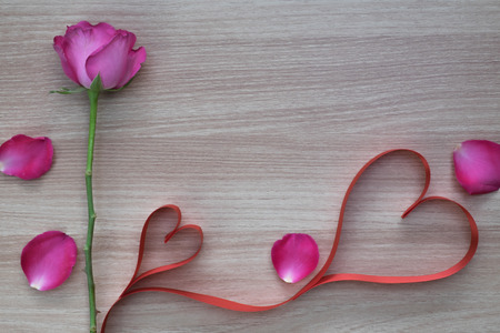 Two red heart shape ribbon with pink rose and petals on wooden surface for valentines or marriage proposal with space for textの写真素材