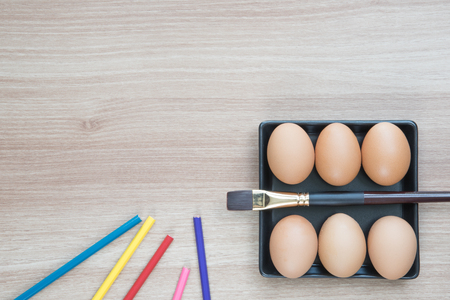 Six eggs in black square plate with a brush and color pencils on wooden surface ready for Easter painting with space for textの写真素材