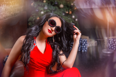 A beautiful, charming girl with sunglasses and a red dress. On the background of a New Year treeの写真素材