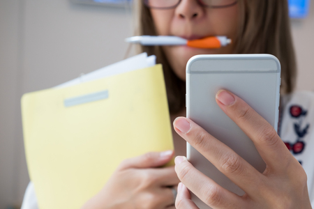 A busy lady holding cellphone and files, and also hold a pen in her mouth.の写真素材