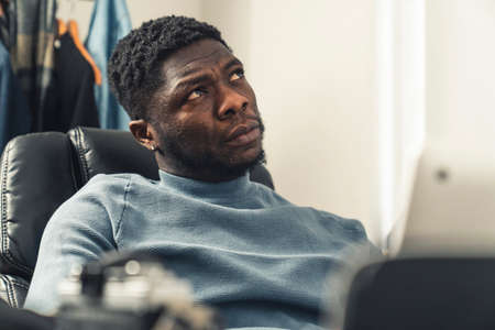 Thinking black man on black chair working on laptop looking up - portrait shot. High quality photoの写真素材