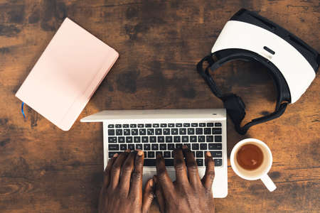Dark wood working desk man writing on computer laptop with notebook VR headset and coffe cup - overtop shot. High quality photoの写真素材