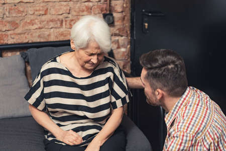 Caucasian grey-haired upset grandma sitting on a sofa supported by her middle-aged son. High quality photoの写真素材