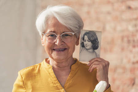 grinning and content grey-haired grandma comparing herself to her old black and white portrait ....