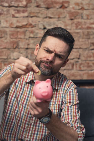 vertical shot of a man holding pink piggy bank and wondering whether to save money or not. High quality photoの写真素材