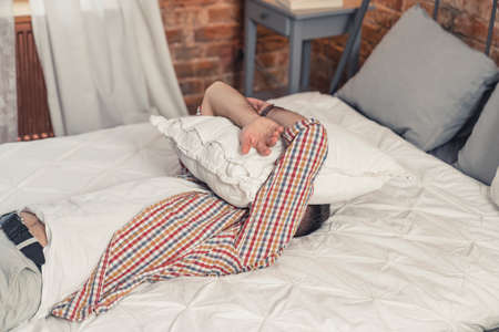 tired caucasian man laying down on his bed and covering his face with white pillow. High quality photoの写真素材