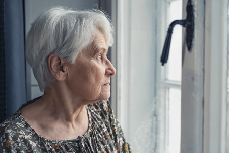 grey-haired sad upset elderly woman in her 60s looking out of the window medium close up indoor shot natural light. High quality photoの写真素材