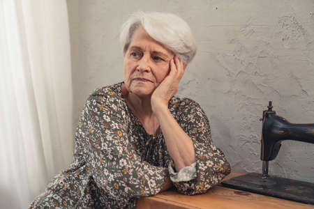 upset caucasian pensioner in her 70s sitting alone next to the sewing machine and propping up her wrinkled chick. High quality photoの写真素材