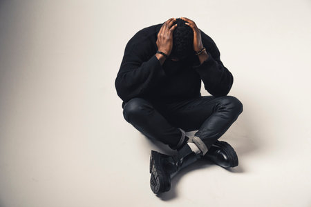 distressed Afroamerican with hands in the back of his head seated white background - full shot. High-quality photoの写真素材