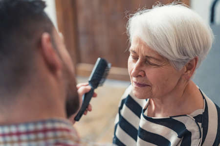 sad retired grandmother closes her eyes and her adult grandson uses a comb to fix her grey hair. High quality photoの写真素材