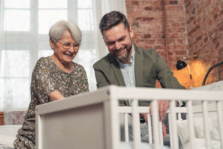 elderly caucasian pensioner grandma and her middle-aged bearded son looking cheerfully at newborn baby in a white crib. High quality photoの写真素材