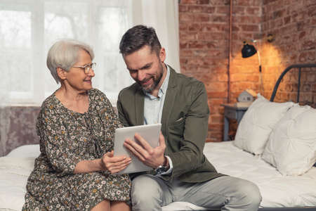 senior grey-haired caucasian lady enjoying her time and watching photos on her middle-aged sons tablet. High quality photoの写真素材