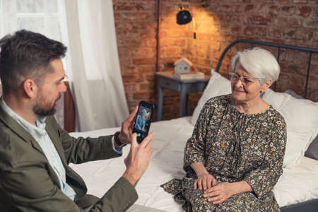 middle-age caucasian entrepreneur taking photo of his elderly grey-haired mother with his smartphone . High quality photoの写真素材